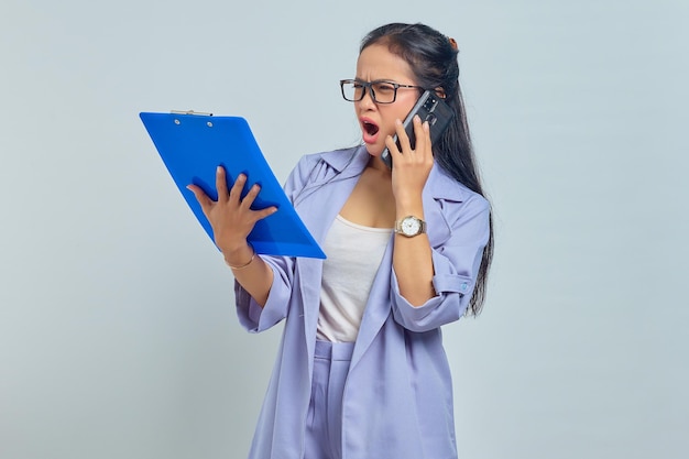 Portrait d'une jeune femme asiatique surprise parlant sur mobile et regardant la liste des tâches dans le dossier de documents isolé sur fond violet