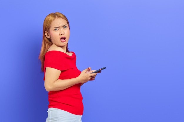 Portrait de jeune femme asiatique surprise en robe rouge, cheveux blonds courts à l'aide d'un téléphone portable isolé sur fond violet