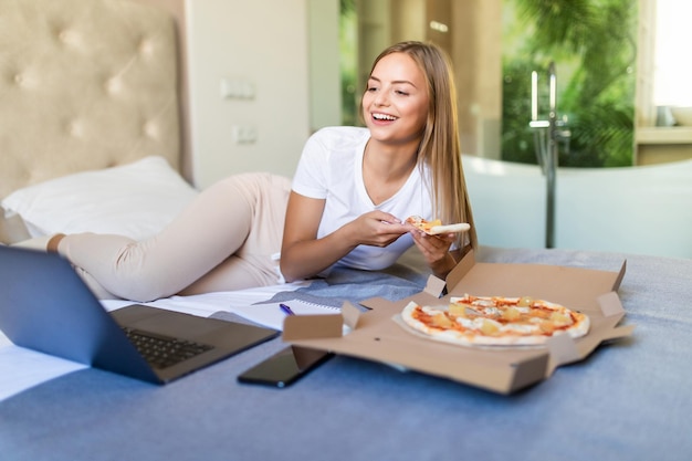 Portrait de jeune femme assise sur un lit avec une boîte de pizza, mordant un morceau de pizza