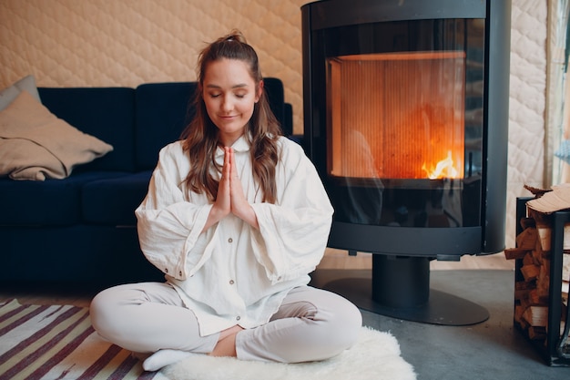 Portrait de jeune femme assise en position du lotus à l'intérieur avec cheminée yoga et méditation zen lik...