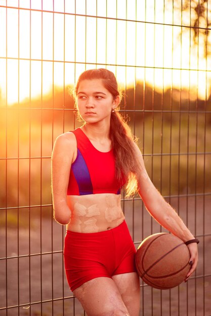 Portrait jeune femme athlète avec un bras amputé et des brûlures sur son corps Elle tient dans les mains le ballon de basket après l'entraînement en plein air au coucher du soleil