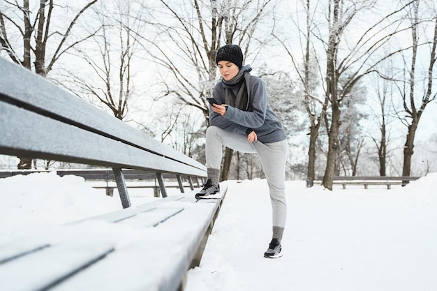 Portrait d'une jeune femme athlète utilisant un smartphone pendant son entraînement d'hiver dans le parc enneigé de la ville