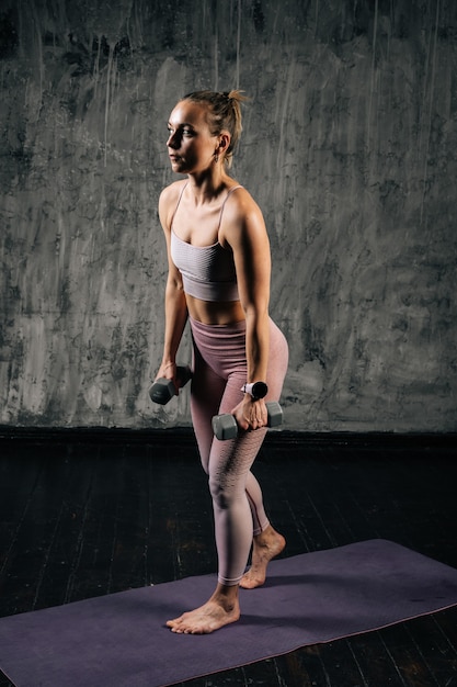 Portrait de jeune femme athlétique musclée avec un beau corps parfait portant des vêtements de sport tenant des haltères et faisant des squats. Femme de remise en forme caucasienne qui pose en studio avec un fond gris foncé.