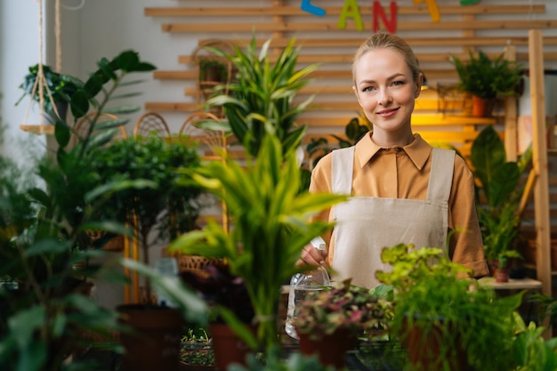 Portrait d'une jeune femme attirante en tablier pulvérisant de l'eau sur des plantes d'intérieur dans des pots de fleurs par pulvérisateur regardant la caméra