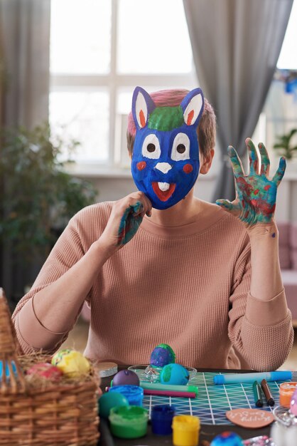 Portrait de jeune femme au masque peint de lapin se présentant à la caméra alors qu'il était assis à la table avec des œufs peints
