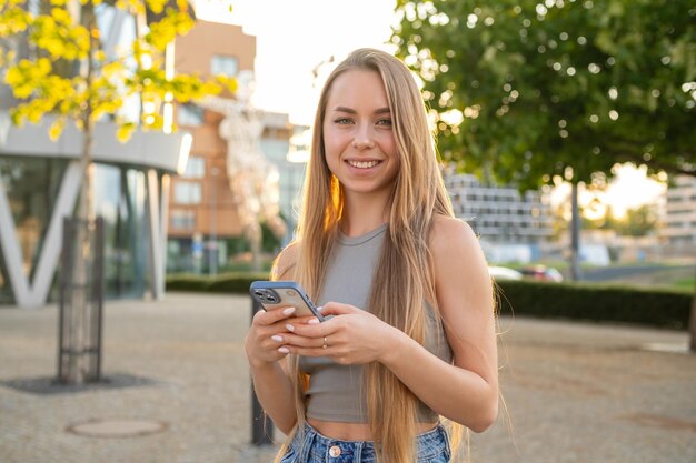 Portrait d'une jeune femme aux cheveux blonds avec un beau sourire debout dans la rue et utilisant un mobile