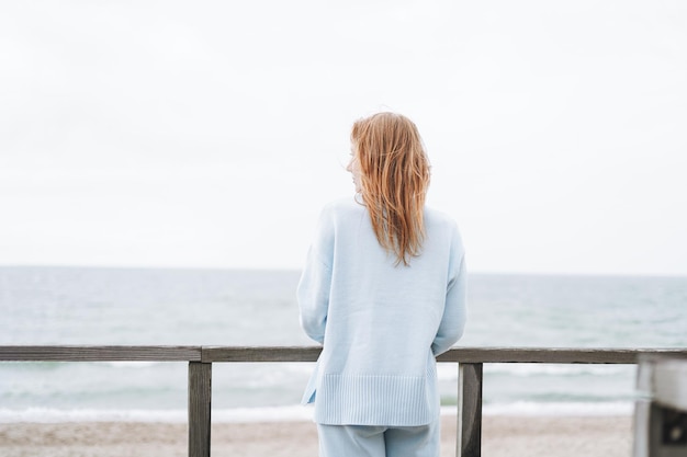 Photo portrait de jeune femme aux cheveux rouges en pull bleu clair sur la plage de sable au bord de la mer dans la tempête