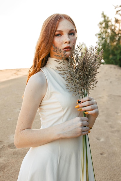 Portrait de jeune femme aux cheveux roux avec des taches de rousseur debout avec de l'herbe sur la plage de sable