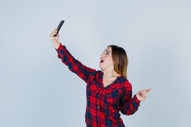 Portrait de jeune femme belle prenant selfie, pointant vers la droite en chemise décontractée et regardant joyeuse vue de face