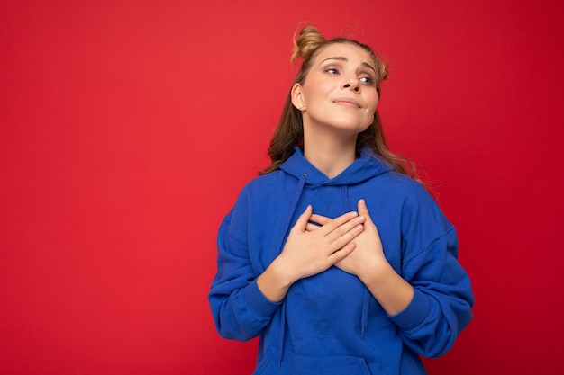 Portrait de jeune femme blonde assez mignonne positive positive avec deux cornes avec des émotions sincères portant des vêtements élégants isolés sur fond rouge avec un espace libre et tenant les mains sur la poitrine.