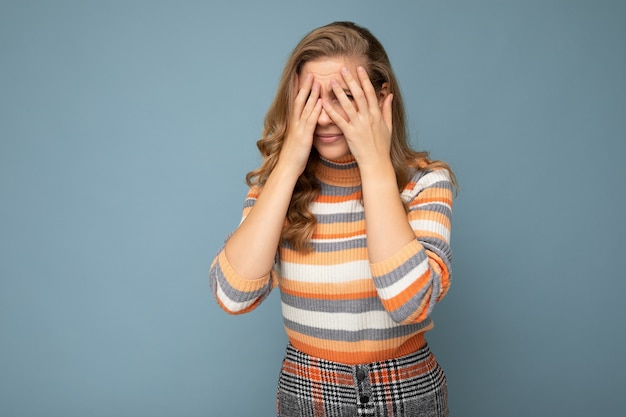 Portrait de jeune femme blonde belle bouclée avec des émotions sincères portant pull rayé isolé sur fond bleu avec un espace vide et couvrant les yeux.