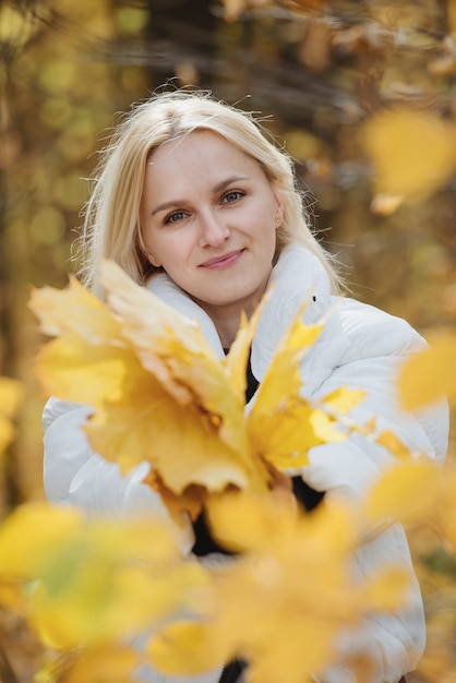 Portrait d'une jeune femme blonde dans la forêt d'automne, avec un bouquet de feuilles jaunes dans ses mains.