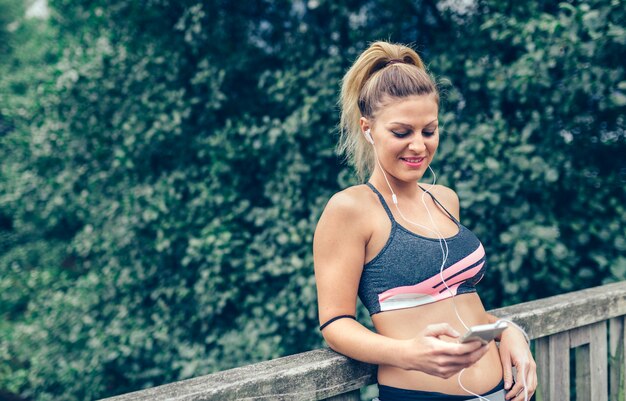 Portrait d'une jeune femme blonde heureuse en vêtements de sport avec des écouteurs regardant son smartphone après une formation à l'extérieur