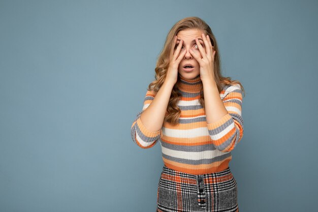 Portrait de jeune femme blonde séduisante bouclée avec des émotions sincères portant un maillot rayé isolé sur fond bleu avec un espace vide et couvrant les yeux.