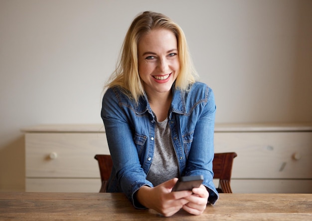 Portrait d'une jeune femme blonde souriante assise à table avec un téléphone portable