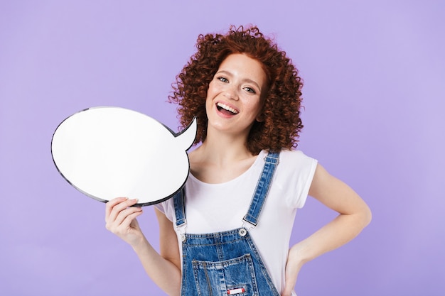 Portrait de jeune femme bouclée rousse des années 20 souriant tout en tenant une bulle de pensée vide isolée sur un mur violet