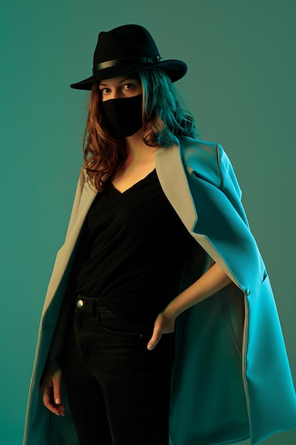 Portrait d'une jeune femme branchée en manteau, chapeau et masque de protection sur fond vert