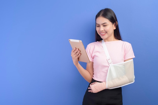 Portrait d'une jeune femme avec un bras blessé dans une écharpe à l'aide d'une tablette sur fond bleu en studio, concept d'assurance et de soins de santé