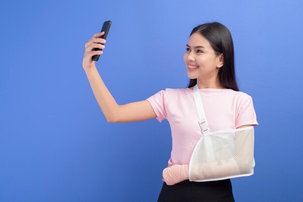Un portrait de jeune femme avec un bras blessé dans une écharpe à l'aide d'un téléphone intelligent sur fond bleu en studio, concept d'assurance et de soins de santé