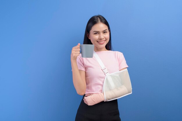 Un portrait de jeune femme avec un bras blessé dans une écharpe ayant une tasse de café sur fond bleu en studio, concept d'assurance et de soins de santé