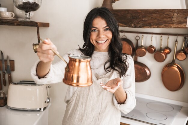 Portrait de jeune femme brune 30s portant un tablier debout avec de la vaisselle à la maison pendant la cuisson dans la cuisine