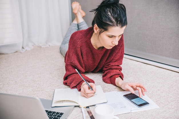 Portrait de jeune femme brune à l'aide d'un ordinateur portable gérant le budget domestique à la maison en prenant des notes avec un crayon assis sur le sol avec une calculatrice de documents d'ordinateur portable ouvert