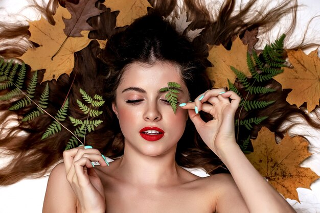 Portrait d'une jeune femme brune sur fond blanc avec une fougère verte et des feuilles jaunes autour de sa tête