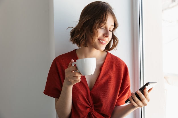 Portrait D'une Jeune Femme Brune Vêtue De Vêtements Décontractés, Buvant Du Café Et Utilisant Un Smartphone Assis Au-dessus De La Fenêtre à La Maison