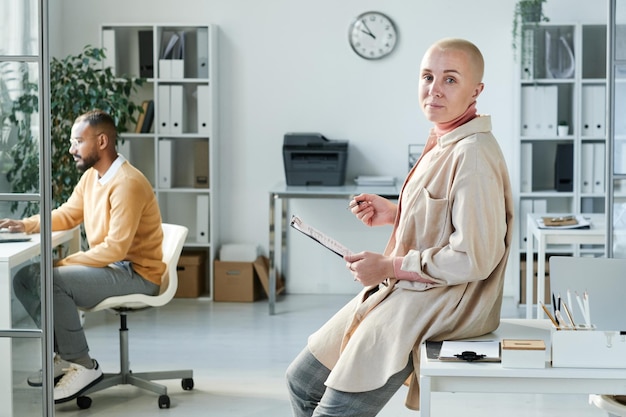 Portrait d'une jeune femme de bureau confiante en chemise longue appuyée sur une table et travaillant avec un document