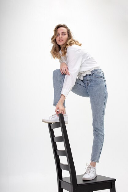 Portrait de jeune femme caucasienne aux longs cheveux bouclés en chemise, jeans, isolé sur fond blanc studio debout sur une chaise.