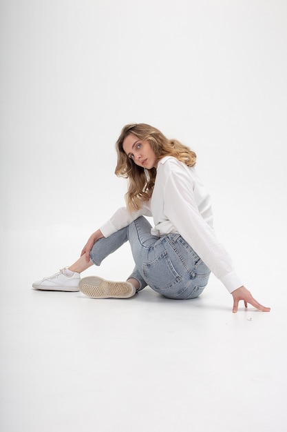 Portrait de jeune femme caucasienne pensive posant en chemise et jeans, assis sur le sol blanc du studio