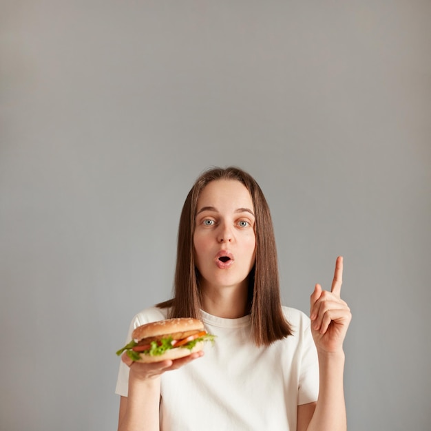 Portrait de jeune femme caucasienne tenant un hamburger isolé sur fond gris dans l'intention de réaliser la solution tout en levant un doigt vers le haut de l'espace de copie pour la publicité