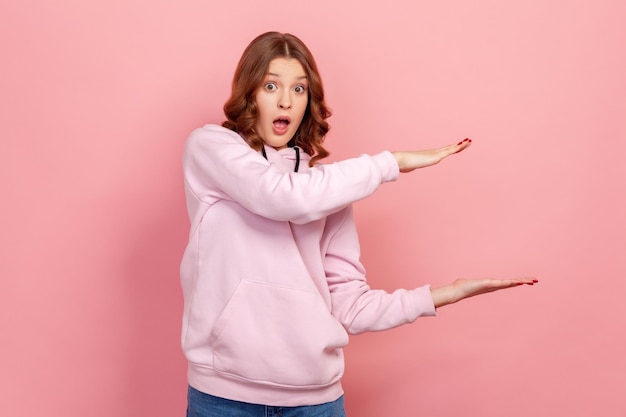 Portrait de jeune femme choquée aux cheveux bouclés en sweat à capuche pointant les bras vers l'espace de copie en regardant la caméra avec la bouche ouverte et les grands yeux discount Prise de vue en studio intérieure isolée sur fond rose