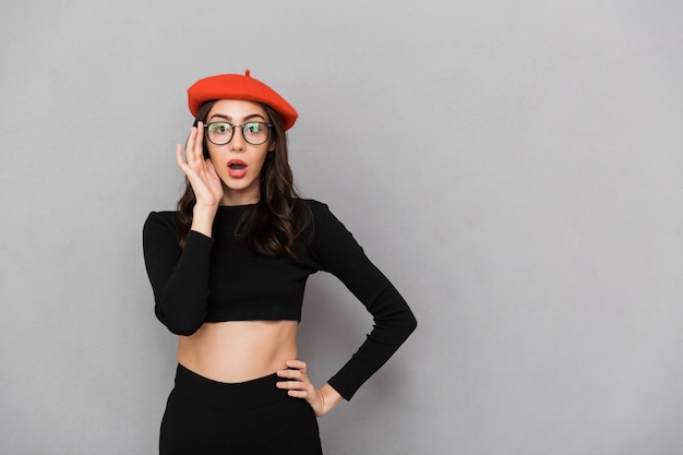 Portrait d'une jeune femme choquée, habillée en chapeau et lunettes debout sur fond gris, regardant la caméra