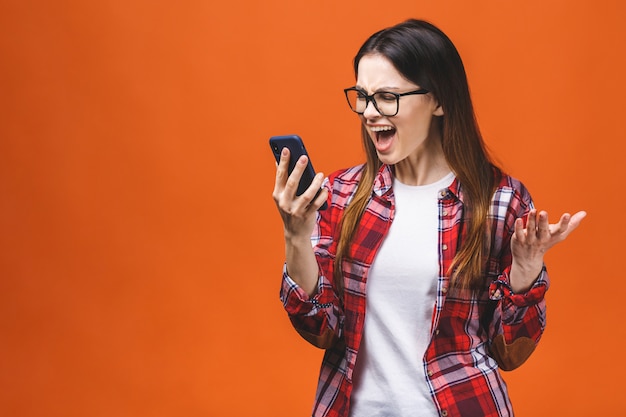 Portrait d'une jeune femme en colère criant sur un téléphone mobile, isolé sur fond orange.