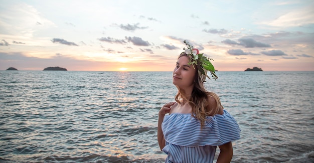 Portrait d'une jeune femme avec une composition de fleurs sur sa tête au bord de l'océan au coucher du soleil.