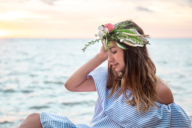 Portrait d'une jeune femme avec une composition de fleurs sur sa tête au bord de l'océan au coucher du soleil.