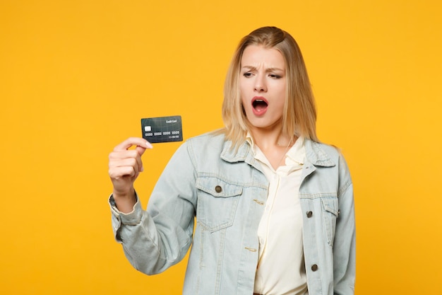 Portrait d'une jeune femme concernée choquée dans des vêtements décontractés en denim tenant une carte bancaire de crédit isolée sur fond de mur orange jaune vif en studio. Concept de style de vie des gens. Maquette de l'espace de copie.