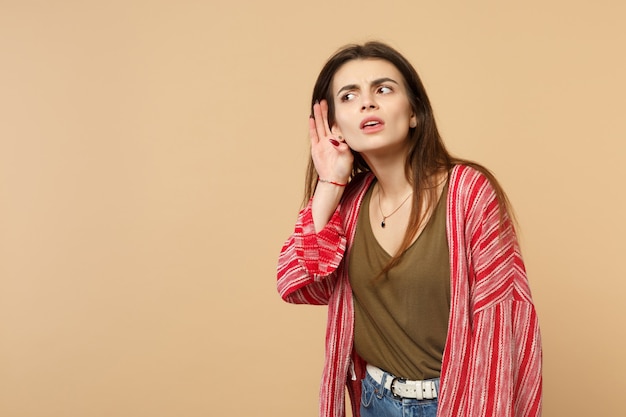 Portrait d'une jeune femme curieuse en vêtements décontractés espionnant avec un geste auditif isolé sur fond de mur beige pastel en studio. Les gens émotions sincères, concept de style de vie. Maquette de l'espace de copie.