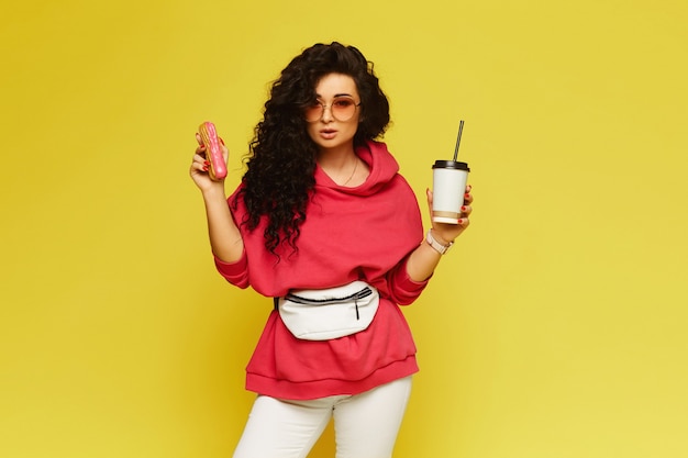 Portrait D'une Jeune Femme Dans Un Sweat à Capuche Rose Tenant Une Tasse De Café En Papier Et éclair Sur Un Mur Jaune, Isolé