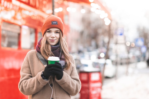 Portrait d'une jeune femme dans des vêtements chauds et des écouteurs, debout sur le fond de la rue, tasse de café en papier dans ses mains