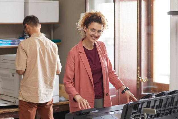 Portrait de jeune femme debout à la machine imprimée et souriant à la caméra avec son collègue travaillant