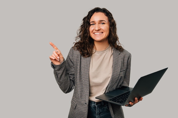 Portrait de jeune femme décontractée tenant un ordinateur portable et pointant vers l'espace de copie sur fond gris