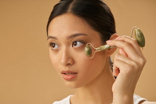 Portrait d'une jeune femme détendue regardant ailleurs, utilisant un rouleau de jade pour masser son visage, posant isolée sur fond beige