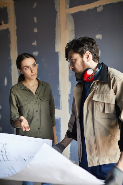 Portrait de jeune femme discutant des plans d'étage avec l'entrepreneur en bâtiment en se tenant debout contre un mur sec en construction maison, copiez l'espace