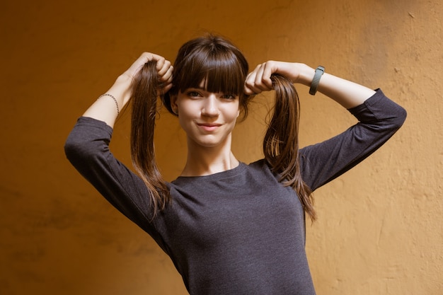 Portrait d'une jeune femme drôle tenant ses cheveux dans ses mains en regardant la caméra contre un mur orange.