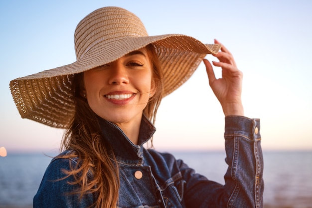 Portrait d'une jeune femme élégante et heureuse dans une veste en jean blanche et un chapeau de paille au bord de l'océan au coucher du soleil, regardant au loin