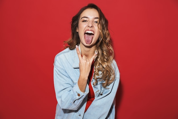 Portrait d'une jeune femme élégante et joyeuse portant une veste en jean debout isolée sur un mur rouge, posant, les cornes vers le haut