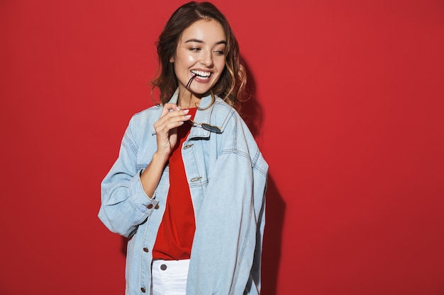 Portrait d'une jeune femme élégante et joyeuse portant une veste en jean et des lunettes de soleil isolées sur un mur rouge, posant