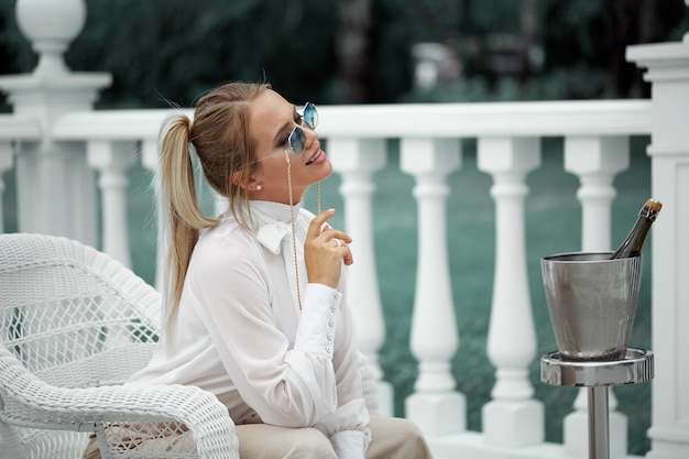 Portrait d'une jeune femme élégante posant dans des lunettes de soleil, assise sur une chaise en osier blanc